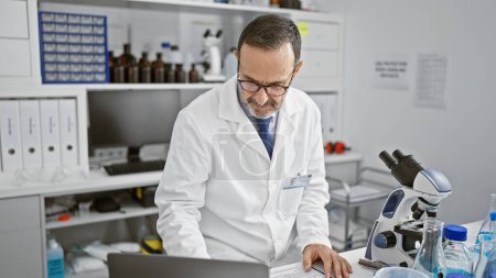 Foto de Hombre serio y enfocado de mediana edad con cabello gris, un científico maduro, absorto en su investigación médica usando un portátil en la mesa de su laboratorio. - Imagen libre de derechos