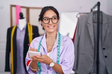 Téléchargez les photos : Jeune belle femme hispanique tailleur souriant écriture confiante sur ordinateur portable au magasin de tailleur - en image libre de droit
