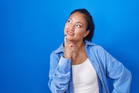 Photo for Asian young woman standing over blue background with hand on chin thinking about question, pensive expression. smiling and thoughtful face. doubt concept. - Royalty Free Image