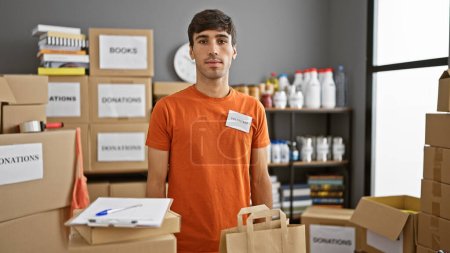 Photo for Serious-faced young hispanic man, a dedicated volunteer, standing at the heart of the charity center, immersed in community service - Royalty Free Image