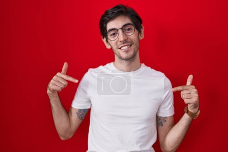 Foto de Joven hombre hispano de pie sobre fondo rojo mirando confiado con sonrisa en la cara, señalándose con los dedos orgullosos y felices. - Imagen libre de derechos