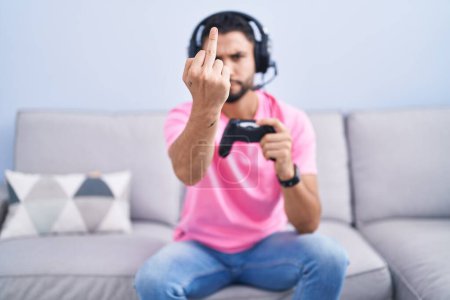 Téléchargez les photos : Hispanic young man playing video game holding controller sitting on the sofa showing middle finger, impolite and rude fuck off expression - en image libre de droit