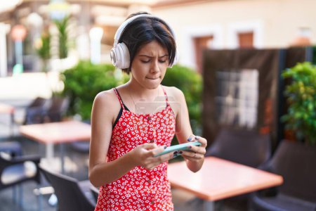 Téléchargez les photos : Adorable fille hispanique jouant à un jeu vidéo avec une expression concentrée dans la rue - en image libre de droit