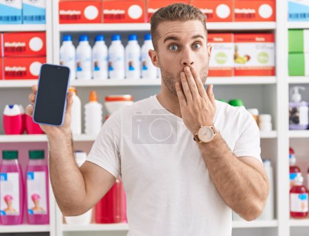 Foto de Joven hombre caucásico que trabaja en la farmacia mostrando la pantalla del teléfono inteligente cubriendo la boca con la mano, sorprendido y asustado por el error. expresión sorprendida - Imagen libre de derechos