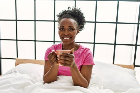 Téléchargez les photos : Femme afro-américaine buvant une tasse de café assise sur le lit dans la chambre - en image libre de droit