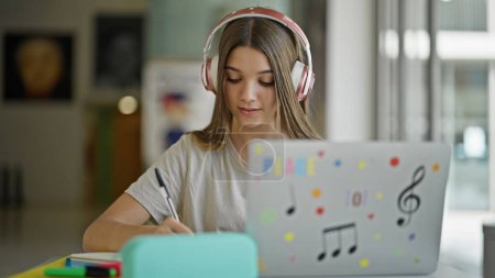 Foto de Joven estudiante hermosa chica usando teléfono inteligente y auriculares tomando notas en la biblioteca - Imagen libre de derechos