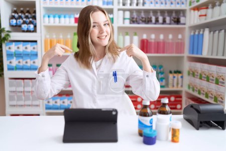 Foto de Mujer caucásica joven que trabaja en farmacia usando tableta que mira confiada con sonrisa en la cara, señalándose con los dedos orgullosos y felices. - Imagen libre de derechos