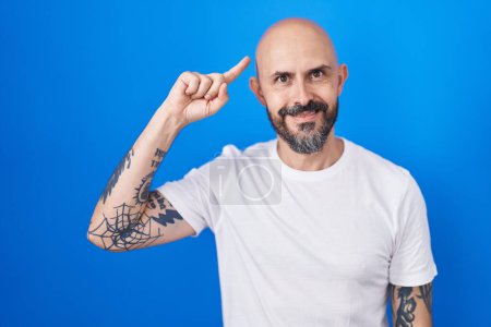 Foto de Hombre hispano con tatuajes de pie sobre fondo azul sonriendo señalando a la cabeza con un dedo, gran idea o pensamiento, buena memoria - Imagen libre de derechos