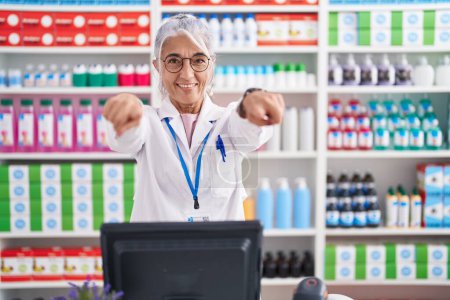Téléchargez les photos : Femme d'âge moyen avec des tatouages travaillant à la pharmacie pharmacie pointant vers vous et la caméra avec les doigts, souriant positif et joyeux - en image libre de droit
