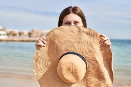 Foto de Joven mujer rubia turista cubriendo la boca con sombrero de verano sonriendo en la playa - Imagen libre de derechos