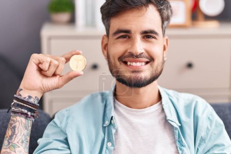 Foto de Joven hombre hispano con tatuajes sosteniendo la moneda criptomoneda atadura mirando positiva y feliz de pie y sonriendo con una sonrisa confiada mostrando los dientes - Imagen libre de derechos