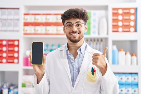Photo for Arab man with beard working at pharmacy drugstore showing smartphone screen smiling happy and positive, thumb up doing excellent and approval sign - Royalty Free Image