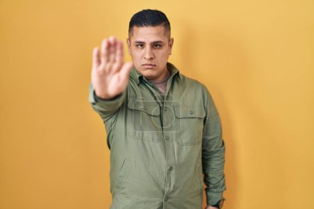 Photo for Hispanic young man standing over yellow background doing stop sing with palm of the hand. warning expression with negative and serious gesture on the face. - Royalty Free Image