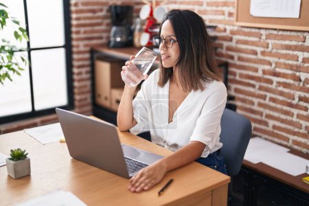 Foto de Young hispanic woman business worker using laptop drinking water at office - Imagen libre de derechos