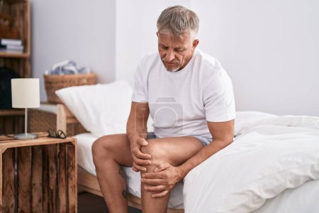 Téléchargez les photos : Middle age grey-haired man suffering for knee injury sitting on bed at bedroom - en image libre de droit