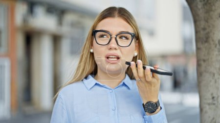 Téléchargez les photos : Jeune femme blonde ouvrière envoyant un message vocal par smartphone travaillant dans la rue - en image libre de droit