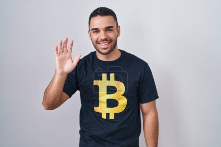 Téléchargez les photos : Jeune homme hispanique portant Bitcoin t-shirt renonçant à dire bonjour heureux et souriant, geste de bienvenue convivial - en image libre de droit