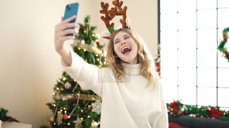 Foto de Mujer rubia joven haciendo selfie por teléfono inteligente de pie junto al árbol de Navidad en casa - Imagen libre de derechos
