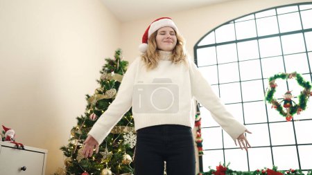 Foto de Joven mujer rubia sonriendo confiada de pie junto al árbol de Navidad en casa - Imagen libre de derechos