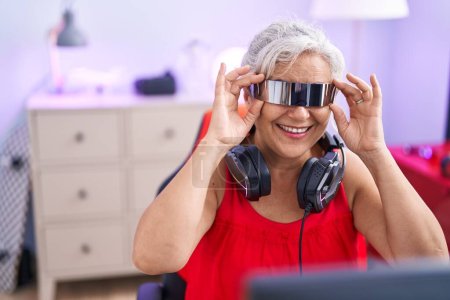 Téléchargez les photos : Femme aux cheveux gris moyen âge streamer jouer à un jeu vidéo en utilisant des lunettes de réalité virtuelle à la salle de jeux - en image libre de droit