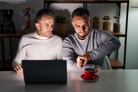 Téléchargez les photos : Couple homosexuel en utilisant ordinateur portable vérifiant l'heure sur la montre-bracelet, détendu et confiant - en image libre de droit