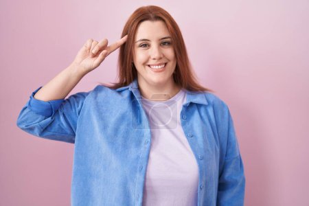 Photo for Young hispanic woman with red hair standing over pink background smiling pointing to head with one finger, great idea or thought, good memory - Royalty Free Image