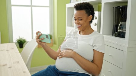Photo for Young pregnant woman watching video on smartphone sitting on table at dinning room - Royalty Free Image