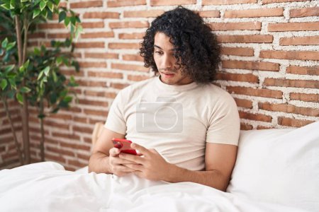 Téléchargez les photos : Homme hispanique avec les cheveux bouclés en utilisant smartphone assis sur le lit effrayé et étonné avec la bouche ouverte pour la surprise, visage incrédule - en image libre de droit
