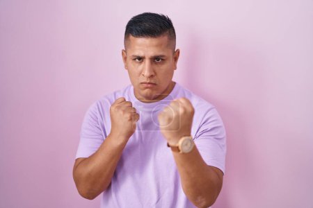 Téléchargez les photos : Young hispanic man standing over pink background ready to fight with fist defense gesture, angry and upset face, afraid of problem - en image libre de droit