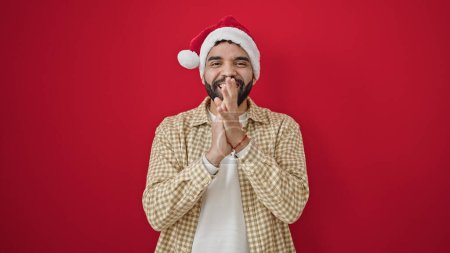 Téléchargez les photos : Jeune homme hispanique portant chapeau de Noël applaudissements mains applaudissements sur fond rouge isolé - en image libre de droit