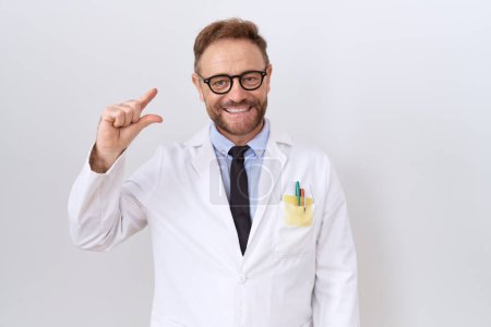 Photo for Middle age doctor man with beard wearing white coat smiling and confident gesturing with hand doing small size sign with fingers looking and the camera. measure concept. - Royalty Free Image