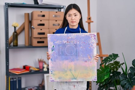 Foto de Mujer joven china sosteniendo lienzo en el estudio de arte escéptico y nervioso, frunciendo el ceño molesto debido a un problema. persona negativa. - Imagen libre de derechos