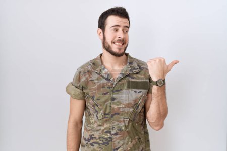 Téléchargez les photos : Jeune homme hispanique portant uniforme de camouflage de l'armée souriant avec le visage heureux regardant et pointant vers le côté avec le pouce vers le haut. - en image libre de droit