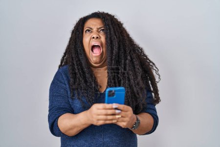 Téléchargez les photos : Plus taille femme hispanique en utilisant le message de dactylographie smartphone en colère et fou hurlant frustré et furieux, criant avec colère levant les yeux. - en image libre de droit