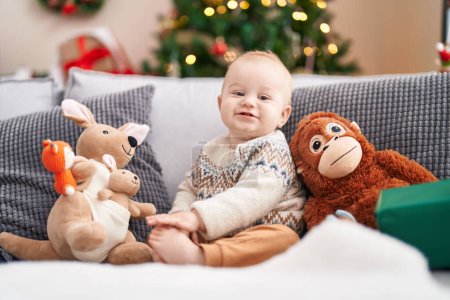 Téléchargez les photos : Adorable bébé caucasien souriant assis sur le canapé par arbre de Noël à la maison - en image libre de droit