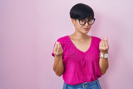 Téléchargez les photos : Jeune femme asiatique avec les cheveux courts debout sur fond rose faisant geste d'argent avec les mains, demandant le paiement du salaire, entreprise millionnaire - en image libre de droit