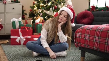 Foto de Mujer rubia joven sentada junto al árbol de Navidad con expresión triste en casa - Imagen libre de derechos