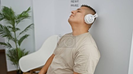 Photo for Young latin man, with a serious expression, relaxed and thoroughly engrossed in the audio of his music, sitting in a waiting room, enjoying his own world indoors. - Royalty Free Image