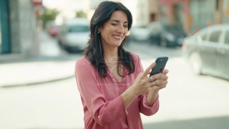 Téléchargez les photos : Moyen Age femme hispanique souriant confiant en utilisant smartphone à la rue - en image libre de droit