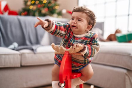 Foto de Adorable niño hispano llorando y jugando con renos meciéndose en el árbol de Navidad en casa - Imagen libre de derechos