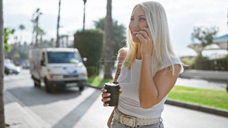 Téléchargez les photos : Une jeune femme blonde confiante rayonne de bonheur, profitant tranquillement d'un café chaud à emporter dans une rue ensoleillée de la ville, tout en parlant et en souriant avec bonheur dans son smartphone dans un portrait de style de vie en plein air - en image libre de droit