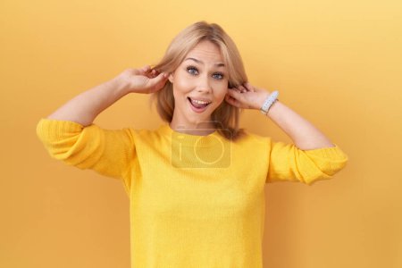 Foto de Mujer caucásica joven con suéter amarillo sonriendo tirando de las orejas con los dedos, gesto divertido. problema de audición - Imagen libre de derechos