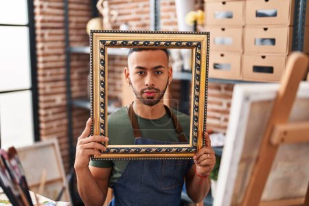 Photo for Young hispanic man sitting at art studio with empty frame relaxed with serious expression on face. simple and natural looking at the camera. - Royalty Free Image