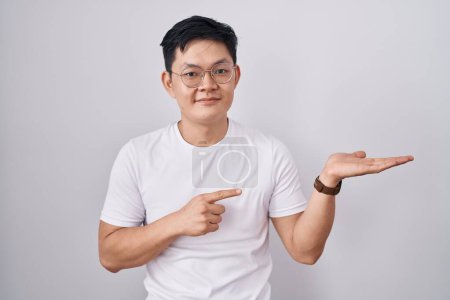 Téléchargez les photos : Jeune homme asiatique debout sur fond blanc étonné et souriant à la caméra tout en présentant avec la main et pointant avec le doigt. - en image libre de droit