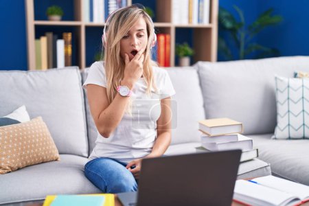 Téléchargez les photos : Jeune femme blonde étudiant à l'aide d'un ordinateur portable à la maison, fascinée par l'incrédulité, la surprise et l'expression étonnante avec les mains sur le menton - en image libre de droit