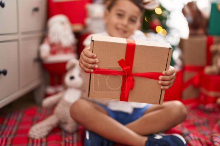 Téléchargez les photos : Adorable bambin hispanique tenant cadeau de Noël assis sur le sol à la maison - en image libre de droit