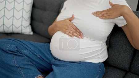 Foto de Mujer embarazada joven examinando el pecho sentado en el sofá en casa - Imagen libre de derechos