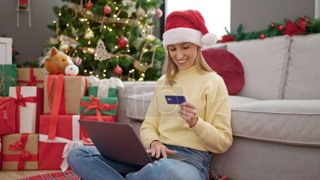 Téléchargez les photos : Jeune femme blonde faisant du shopping avec ordinateur portable et carte de crédit assis sur le sol près de l'arbre de Noël à la maison - en image libre de droit