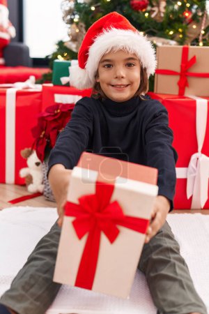 Téléchargez les photos : Adorable garçon hispanique tenant cadeau de Noël assis sur le sol à la maison - en image libre de droit