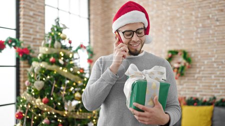 Foto de Hombre hispano con sombrero de navidad sosteniendo regalo hablando por teléfono en casa - Imagen libre de derechos